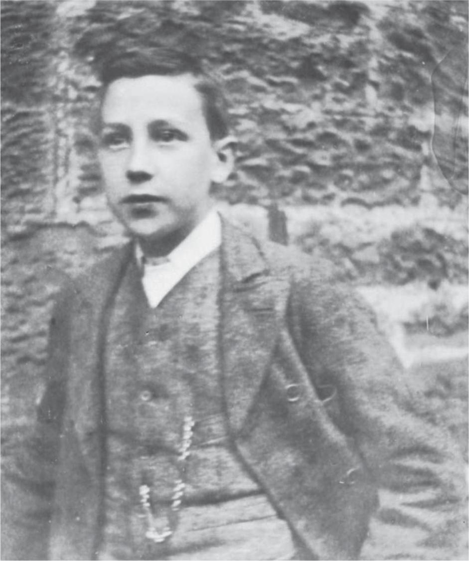 Black and white photograph of a young Alexander Mackie, wearing a jacket and waistcoat, standing in front of a stone wall.