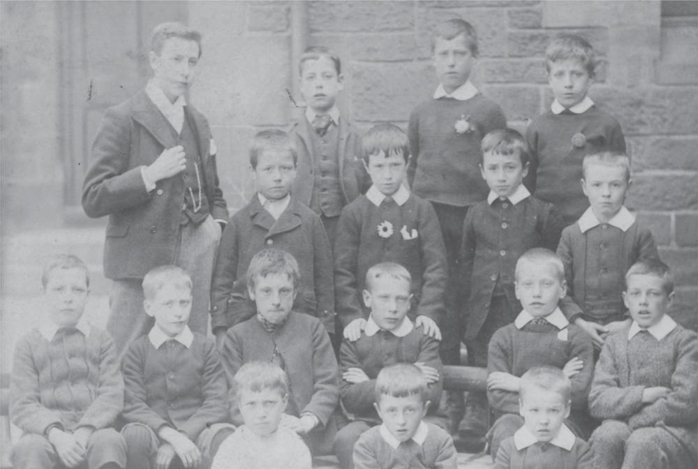 Black and white photograph of a group of children sitting and standing in four rows. A young Alexander Mackie stands on the left side of the photograph.