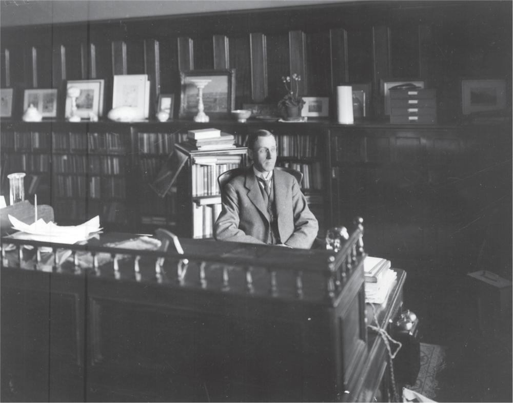 Black and white photograph of Alexander Mackie sitting at a large desk, looking into the distance. The wall behind him is covered with bookcases, filled with books, objects and framed pictures.