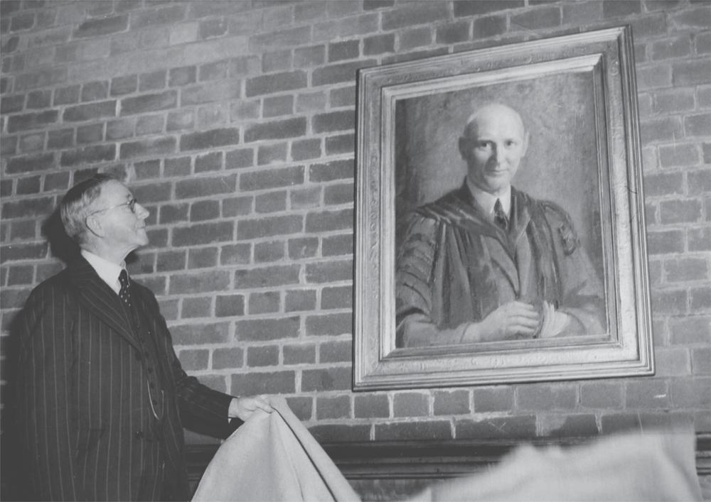 Black and white photograph of Alexander Mackie standing beside a framed portrait of Percy Cole, holding a piece of cloth.