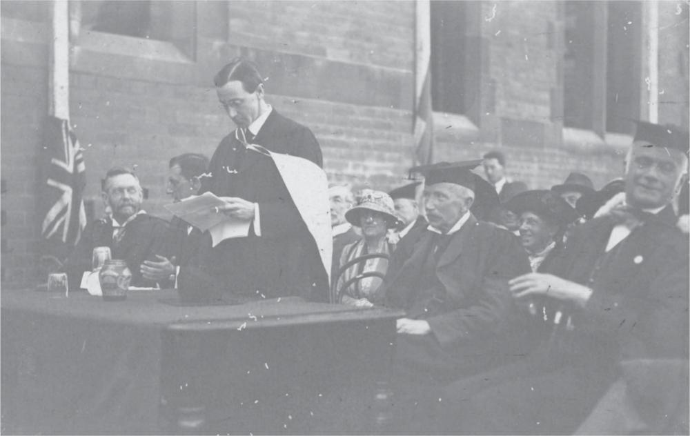 Black and white photograph of four men behind a table. The second man from the left of the picture stands, reading from a piece of paper. The Union Flag hangs beside the table.