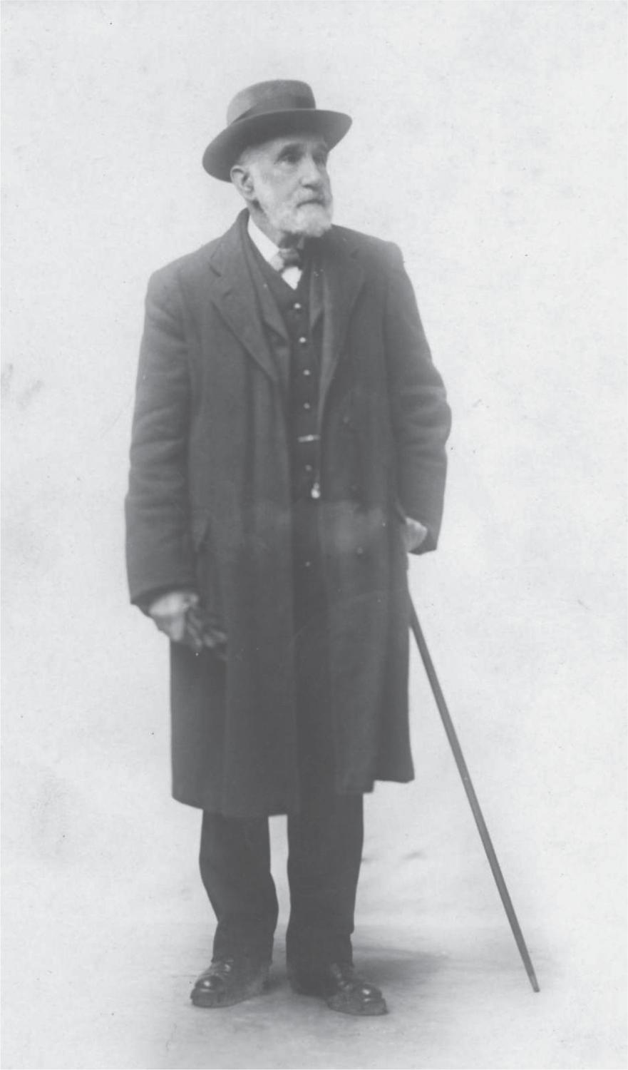Black and white photograph of William Mackie wearing a hat and a long coat and holding a cane.