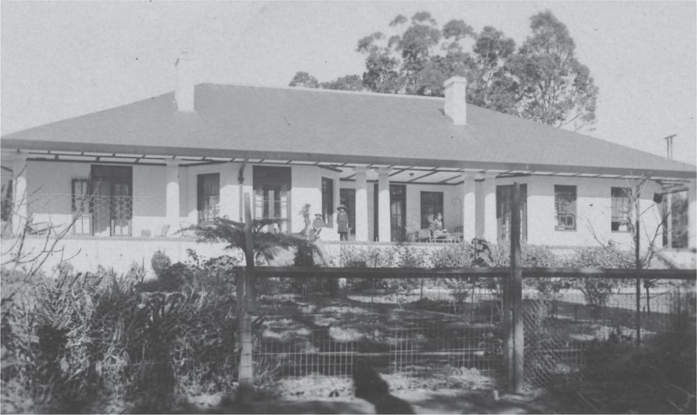 Black and white photograph of a large white house behind a wire fence, with three people on the porch.