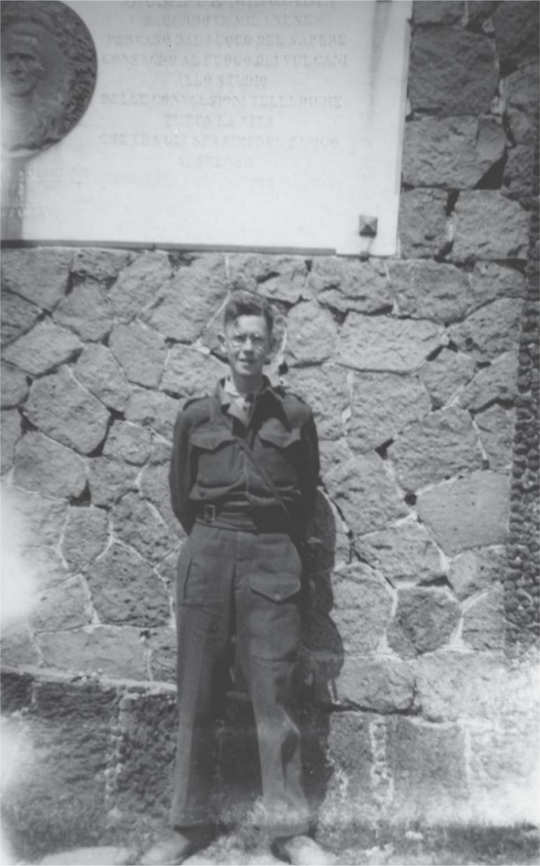 Black and white photograph of John Mackie standing in front of a stone wall. A large, light-coloured, engraved stone sits in the wall above his head.