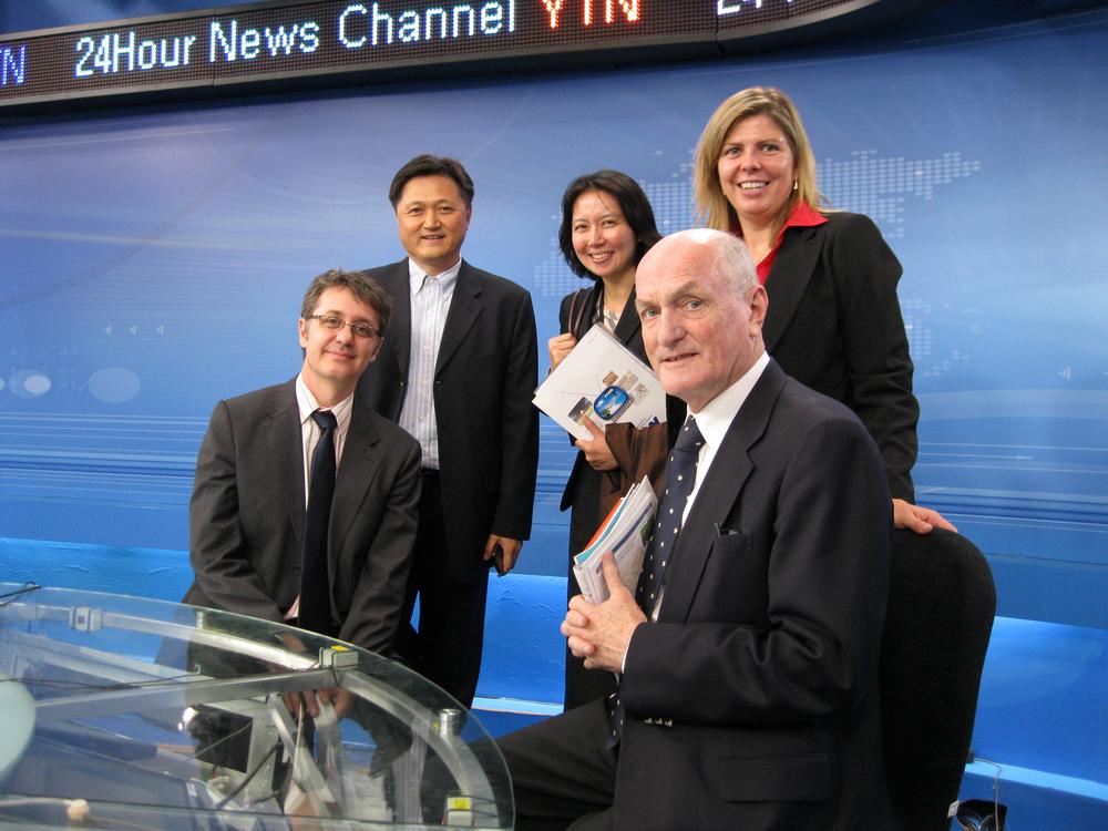 Photograph of five people in a news studio; two are seated and three are standing behind them.