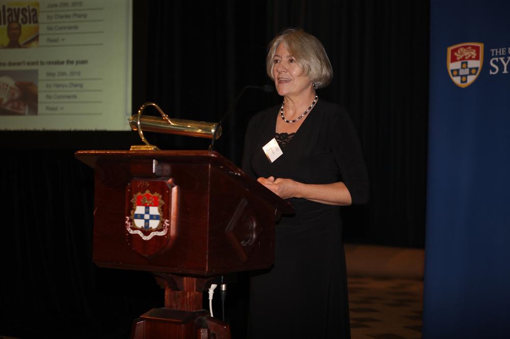 Photograph of a woman with grey hair, dressed in black and standing behind a wooden lectern.
