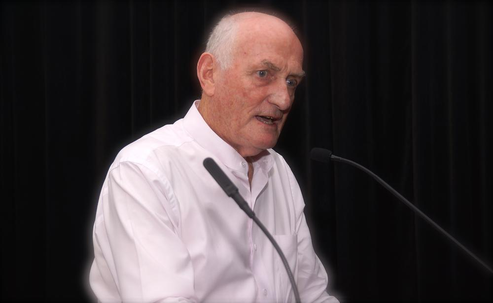 Photograph of a man with grey hair, wearing a white shirt and sitting in front of a microphone.