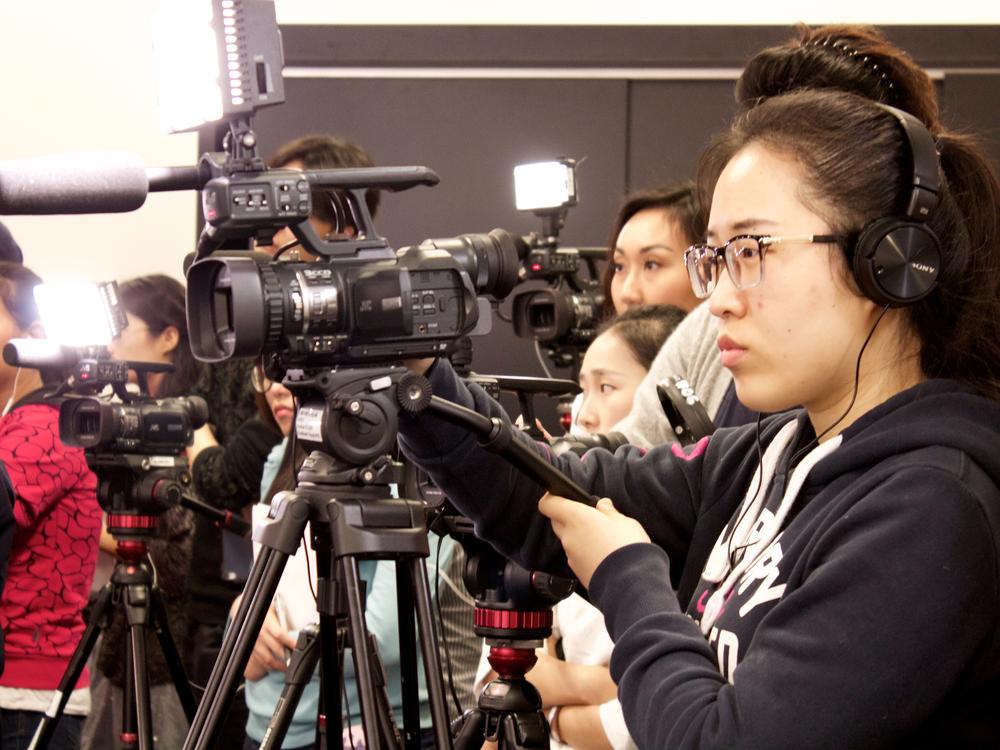 Photograph of a student wearing headphones and operating a camera on a tripod, with a group of people in the background.
