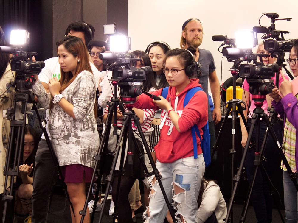 Photograph of five students operating cameras on tripods, with a group of people standing behind them.