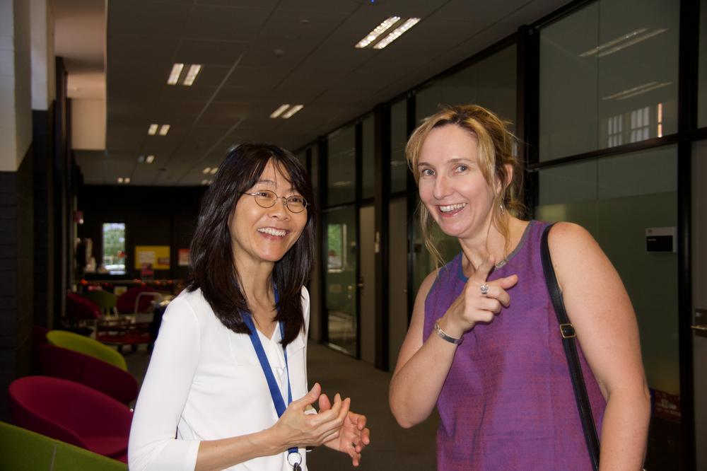 Photograph of two women standing together in a hallway; one is pointing towards the camera.
