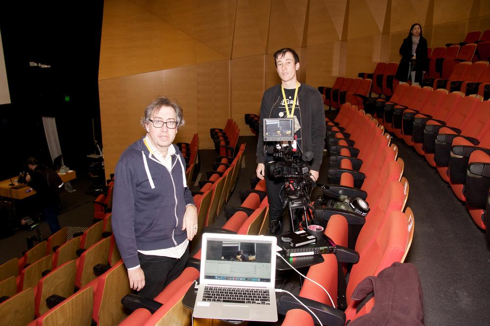 Photograph of two people standing beside a camera on a tripod, among rows of tiered seating.