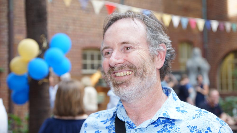 Photograph of a man wearing a blue and white shirt, with balloons and coloured flags visible in the background.