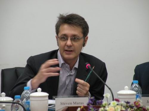 Photograph of a man wearing glasses, a striped shirt and a black jacket, seated at a table with a microphone and a name tag in front of him.