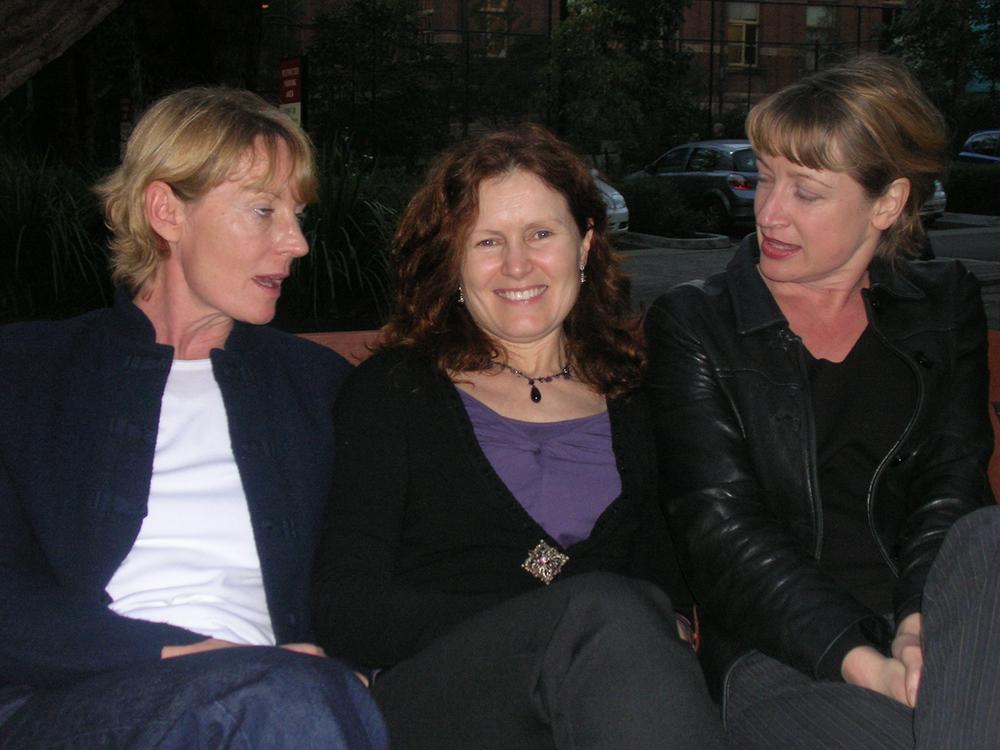 Photograph of three women seated on a couch; two are conversing and one is smiling at the camera.