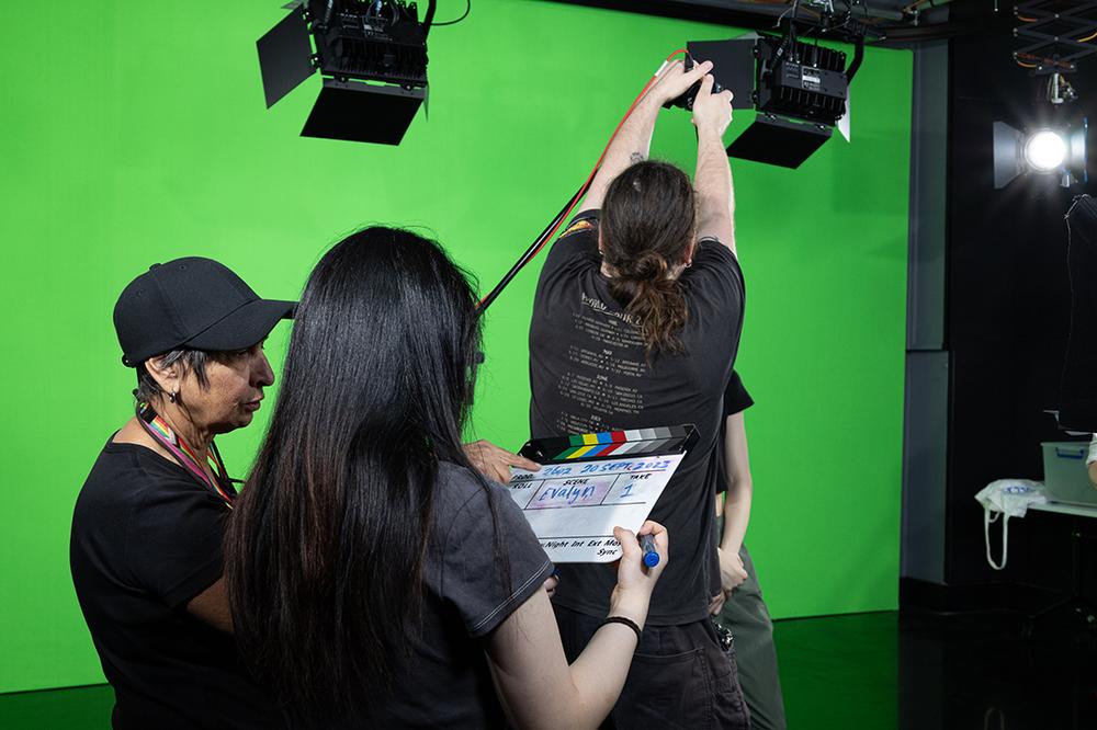 Photograph of two women writing on a clapperboard and a man setting up a microphone, in front of a green screen.