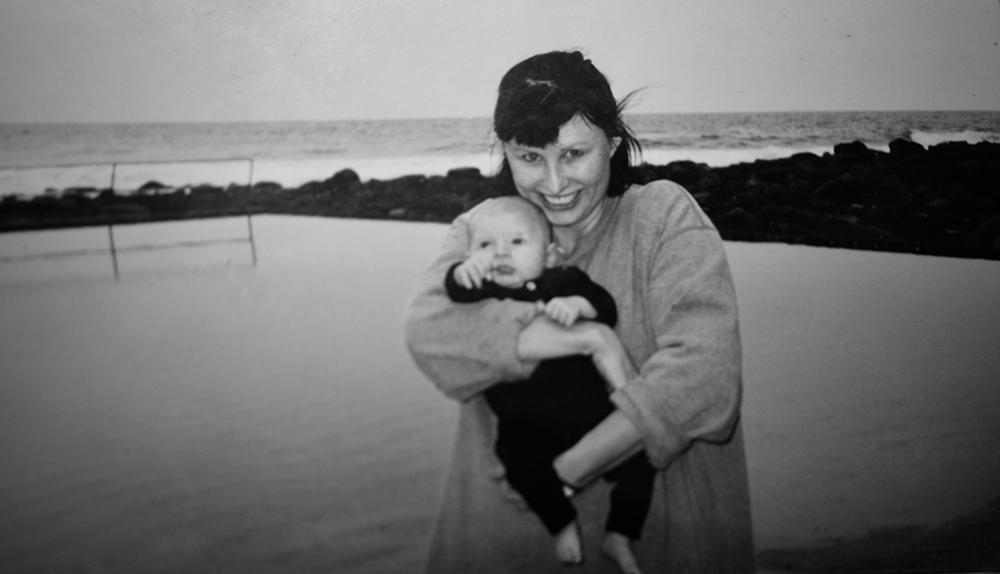 Greyscale photograph of a dark-haired woman holding a young baby, standing beside a pool with the ocean in the background.