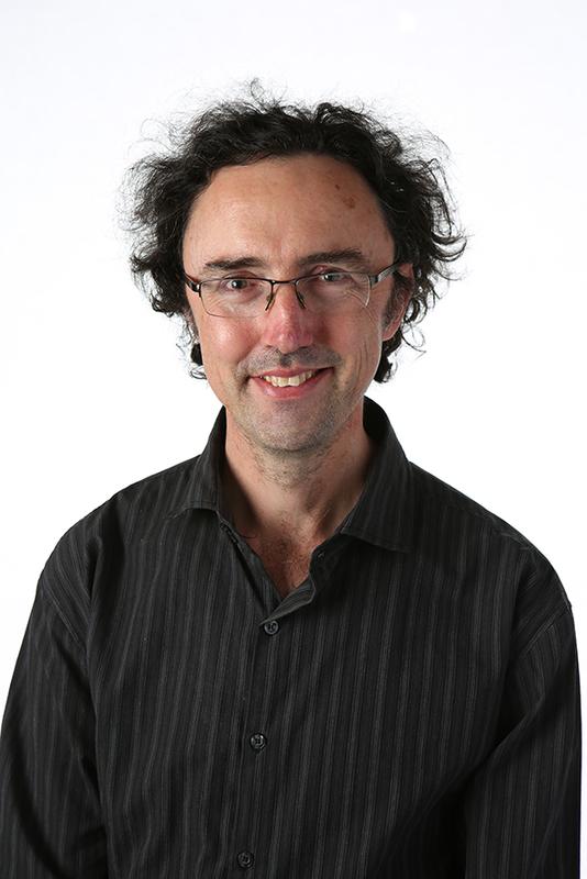 Photograph of a man with dark hair, wearing glasses and a black and grey striped shirt.
