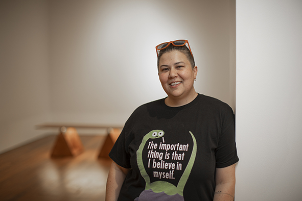 Photograph of a woman with short dark hair, leaning against a wall and wearing a black t-shirt that says 'The important thing is that I believe in myself'.