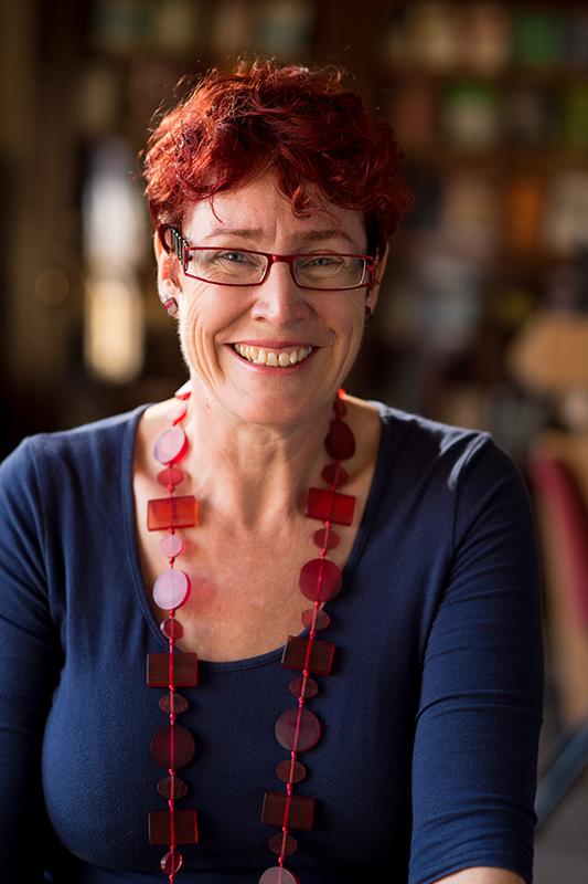 Photograph of a woman with red hair, wearing glasses, a blue top and a beaded necklace.
