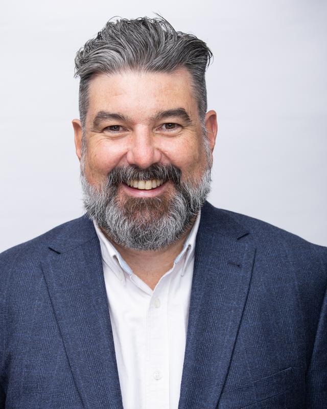 Photograph of a man with dark hair and a beard, wearing a white shirt and a blue jacket.