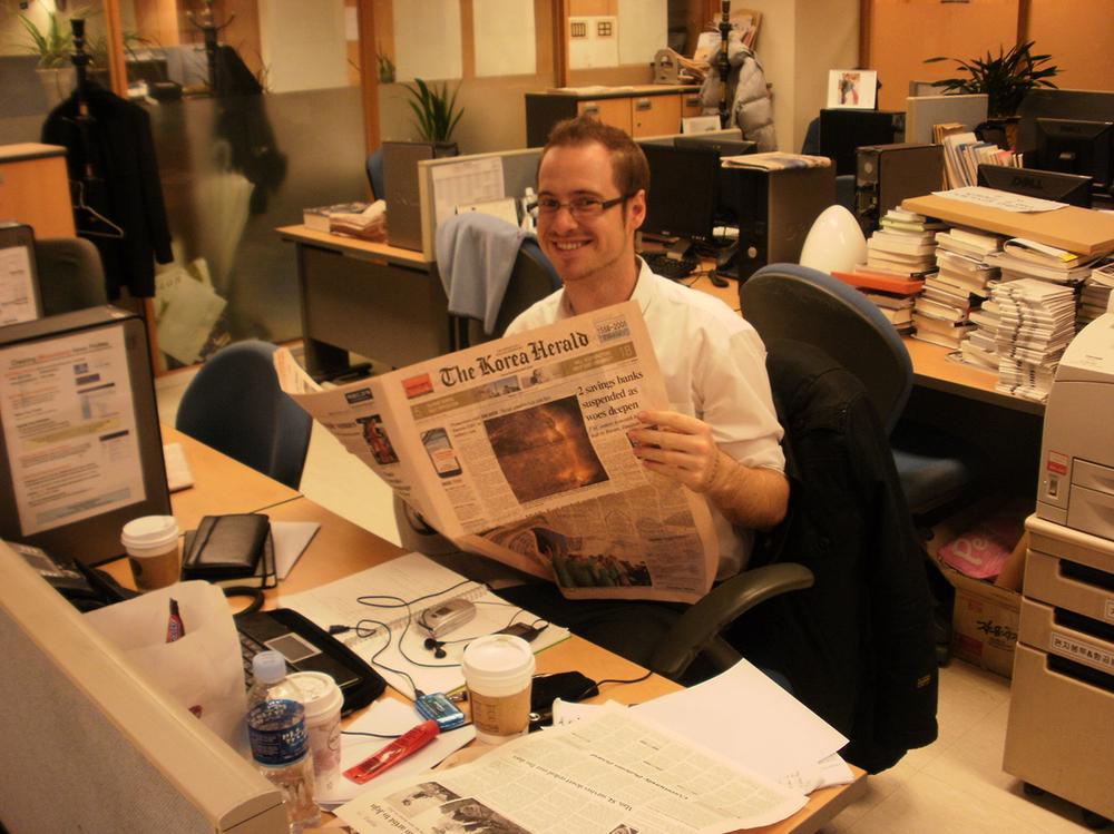 Photograph of a man wearing glasses and a white shirt, sitting at a desk and reading a copy of The Korea Herald.