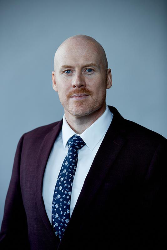 Photograph of a man wearing a white shirt, a patterned blue tie and a burgundy jacket.