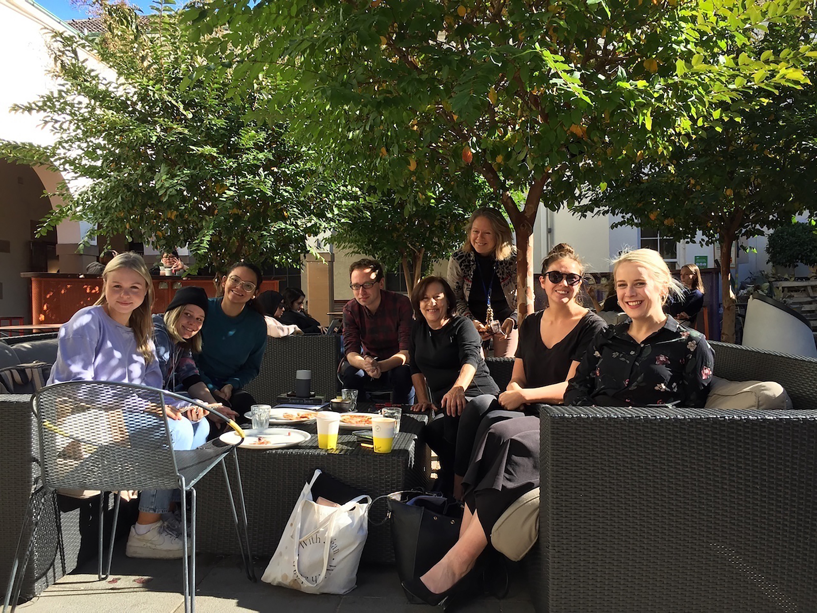 Photograph of eight people sitting around a low table, under a tree.