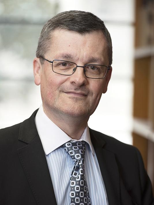 Photograph of a man with dark hair, wearing a striped shirt, a black and white patterned tie and a black jacket.