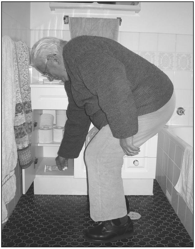 A black and white photograph of an elderly man bending down in a bathroom. The bathroom cabinet is open and the man is reaching to grab something inside. He has a thick jumper and glasses.