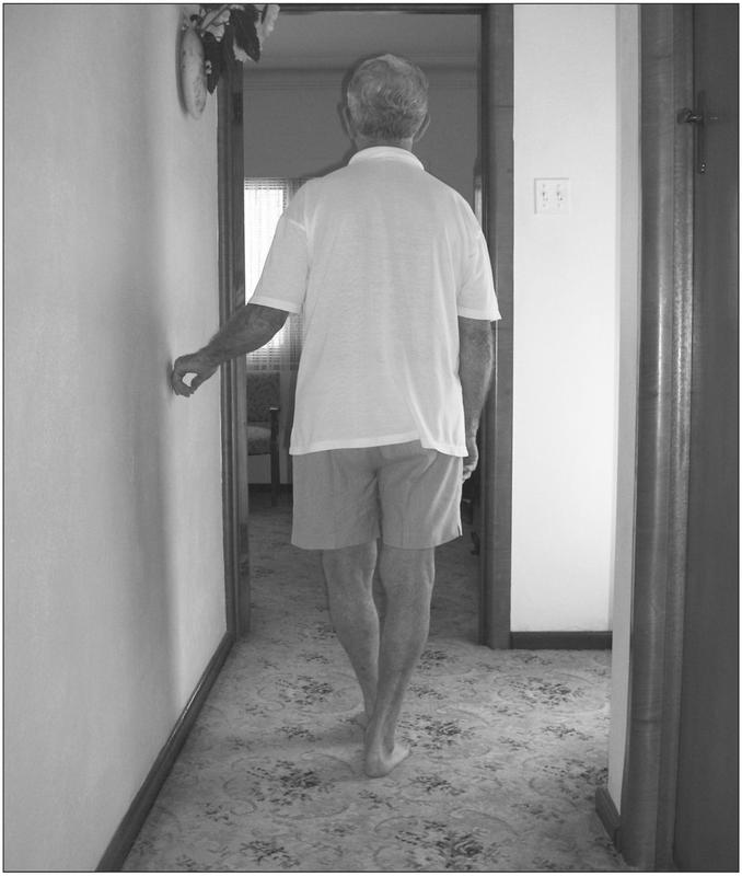 A black and white photograph of an elderly man walking to an open doorway. He has his back to the camera and has a hand resting against the wall. He is wearing a white short-sleeved shirt and shorts.