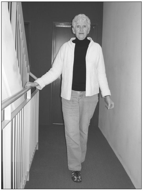 A black and white photograph of an elderly woman walking towards the camera in a hallway. She has one foot in front of the other and has a hand resting on a handrail. She is wearing a jacket with a dark shirt and long pants.