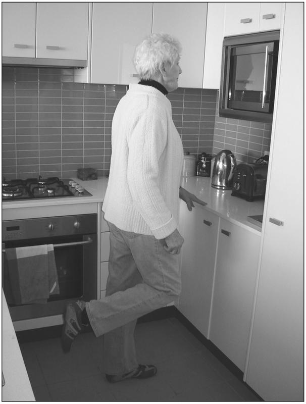 A black and white photograph of an elderly woman balancing on one foot with the other raised behind her. She is in the kitchen and has her fingertips balanced on the countertop.