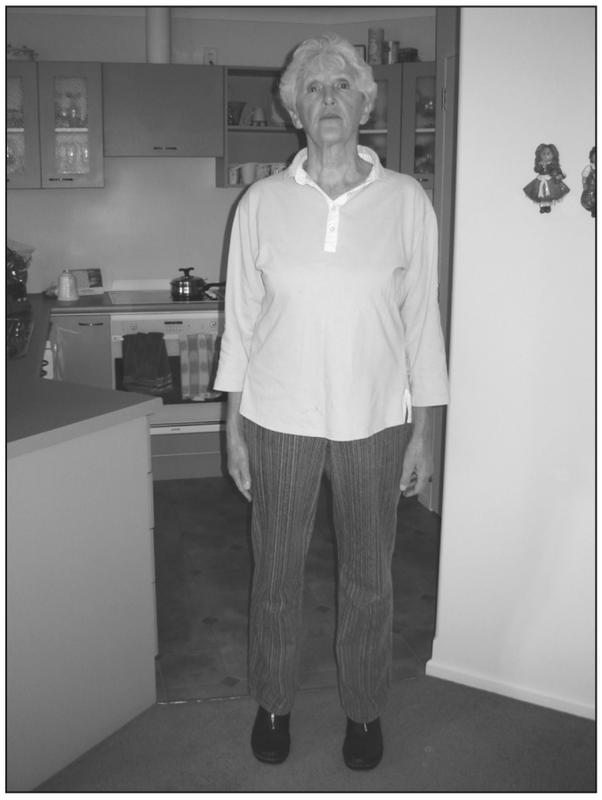 A black and white photograph of an elderly woman standing in a kitchen with her feet apart and her arms resting by her sides. 