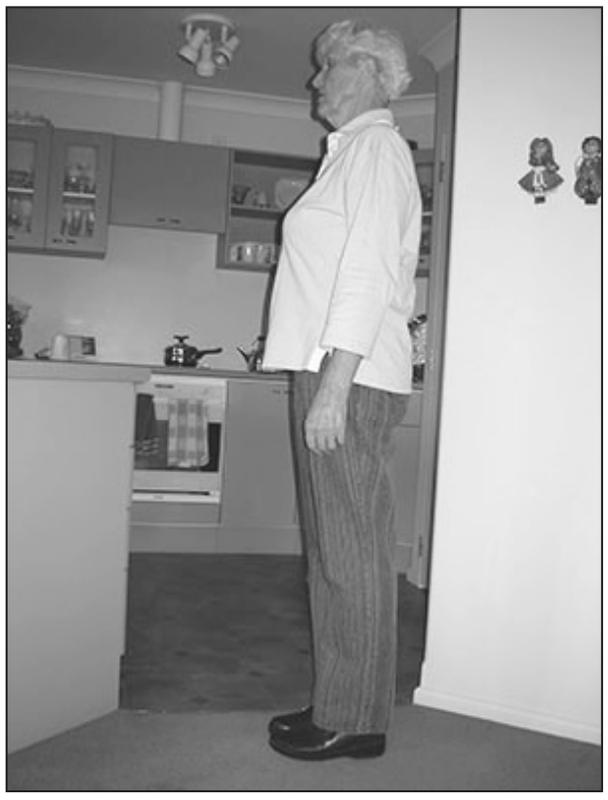 A black and white photograph of an elderly woman standing with her feet apart and arms resting by her side. She is standing by her side so that her half her profile can be seen.