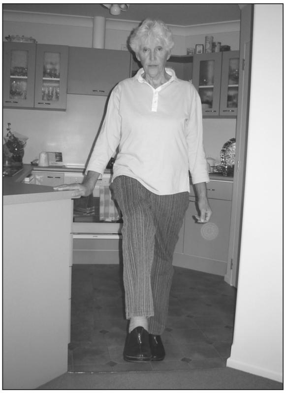 A black and white photograph of an elderly woman facing the camera with one leg slightly raised between the line and carpet. She has one hand clutching the corner of the kitchen countertop.