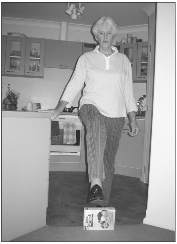 A black and white photograph of an elderly woman facing the camera with one leg raised to step over a muesli box. She has one hand clutching the corner of the kitchen countertop.