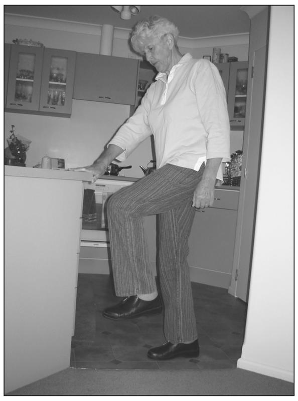 A black and white photograph standing sideways with a leg raised over the line and carpet. She has a hand resting on the corner of a kitchen countertop.