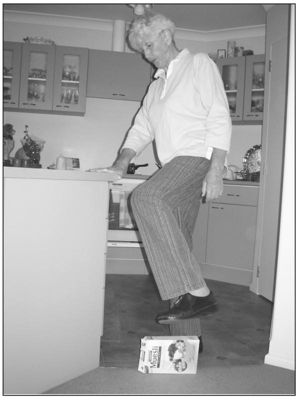 A black and white photograph standing sideways with a leg raised over a muesli box. She has a hand resting on the corner of a kitchen countertop.
