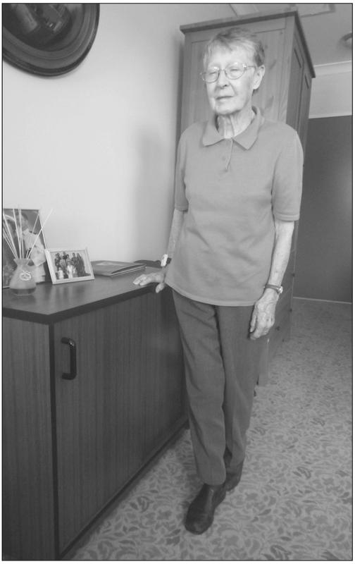 A black and white photograph of an elderly woman standing with her eyes closed in a room with one foot in front of the other and a hand resting on a drawer.