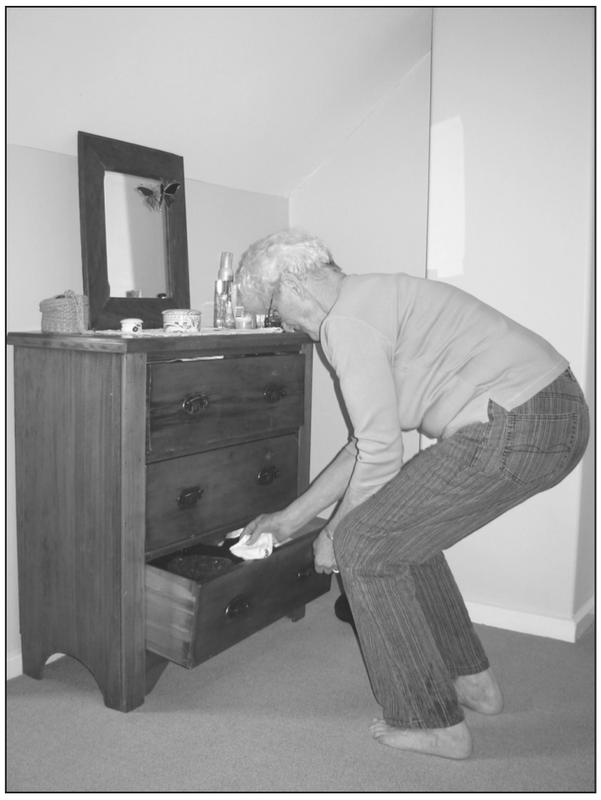 A black and white photograph of an elderly man bending his knees to open the bottom drawer of a cabinet to remove socks. 