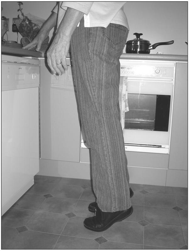 A black and white photograph of an elderly person standing on their toes with the fingertips of their right hand resting on the countertop.