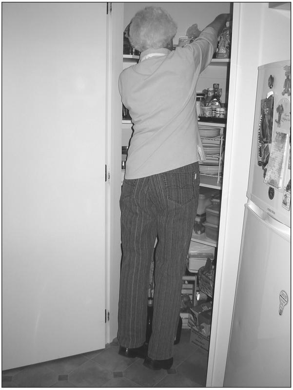 A black and white photograph of an elderly woman standing on her toes with an arm reaching the top shelf of a kitchen cabinet.