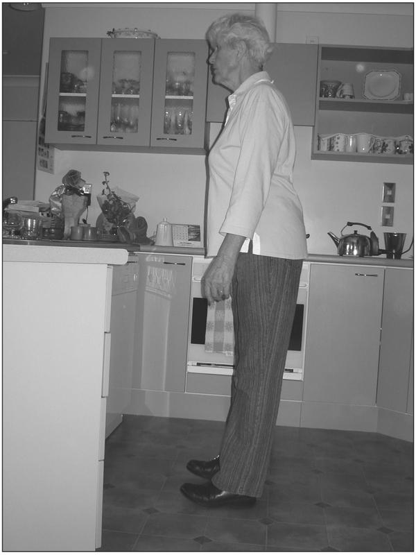 A black and white photograph of an elderly woman standing on her heels in a kitchen. Her hands are by her sides and her chest is leaning slightly forward.