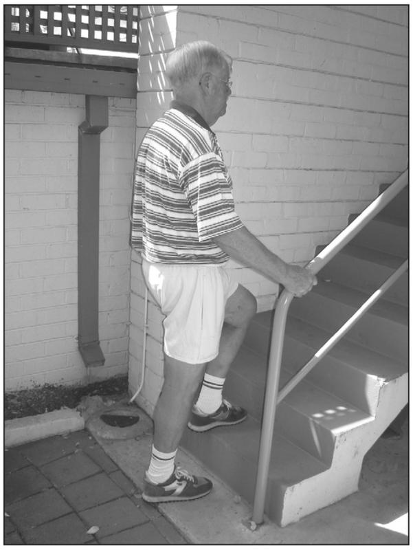 A black and white photograph of an elderly man walking up the stairs with a hand grabbing the handrail. His back is straight and he is wearing a striped shirt and shorts.