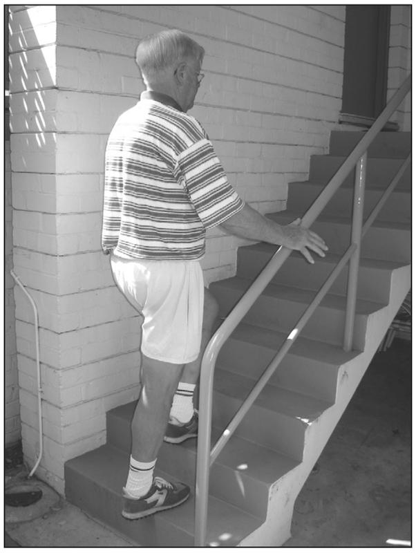 A black and white photograph of an elderly man walking up the stairs with a hand lightly resting on the handrail