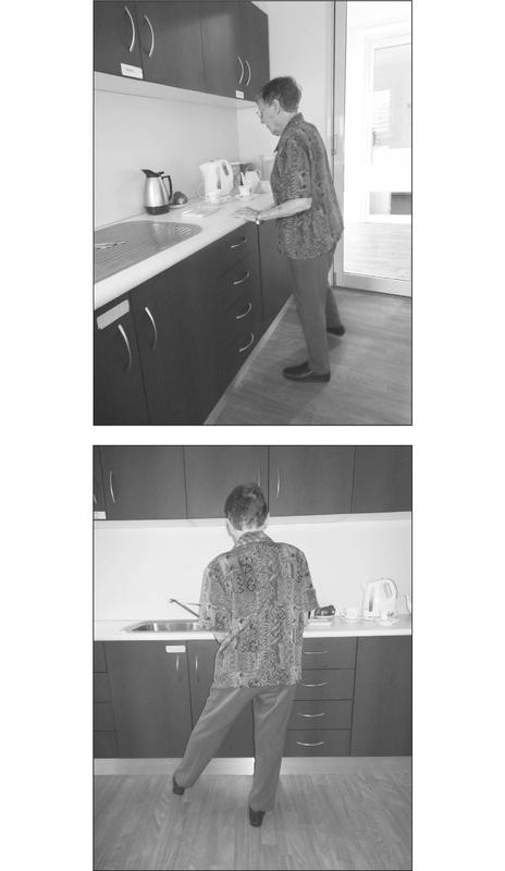 A black and white photograph of an elderly woman standing at a kitchen countertop. She has one leg raised to the side with her hands resting on the counter.