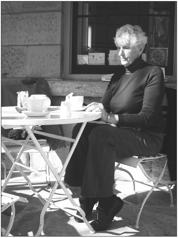 A black and white photograph of an elderly woman sitting at a coffee table with her hands resting on the table. She has her heels up with her weight resting on her toes.