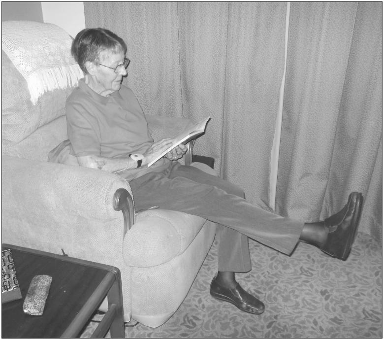 A black and white photograph of an elderly woman sitting in an armchair while reading. She has a leg raised with the other is resting on the ground.
