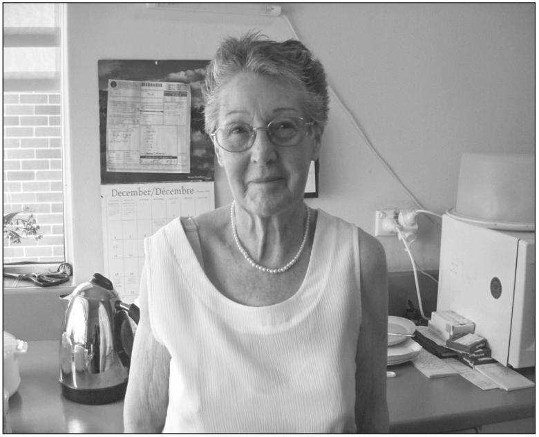 A black and white photograph of an elderly woman smiling at the camera. She is wearing round glasses and a necklace. Her hair is short and she has a sleeveless shirt on.