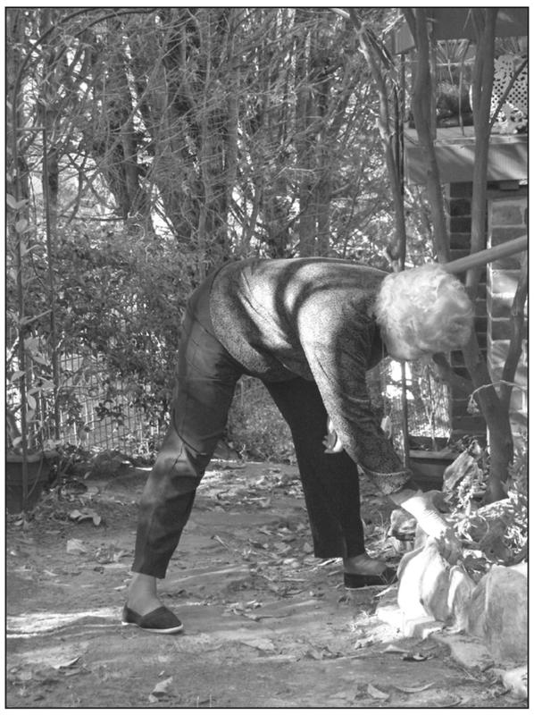 A black and white photograph of an elderly woman bending down with straight legs. She is reaching to pull something from her garden.